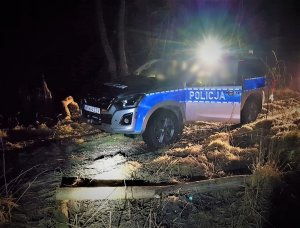 Na tle ciemnego lasu radiowóz marki Isuzu D-MAX z włączonym światłem bocznym